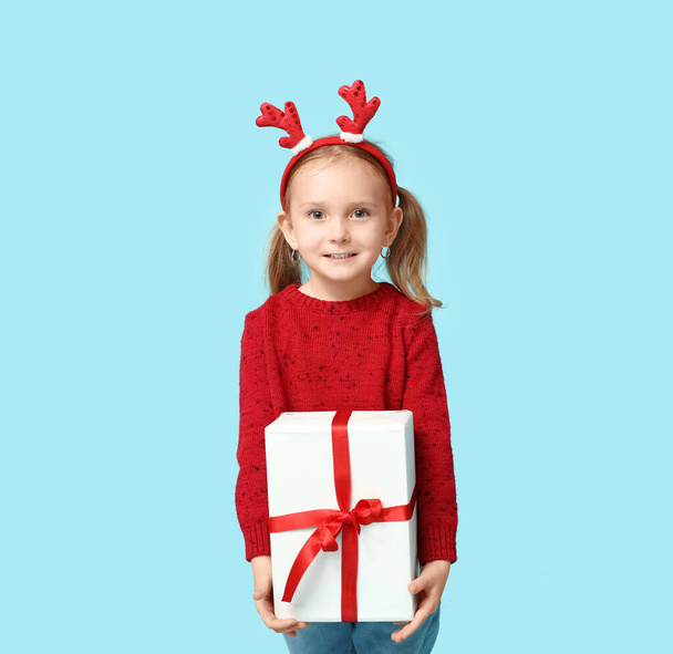 Cute little girl in reindeer horns with Christmas gift on blue background - Photo, Image