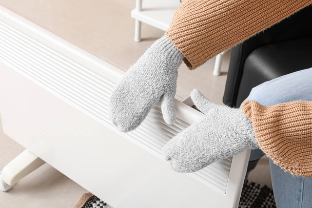 Νεαρή γυναίκα με ζεστά γάντια κοντά σε ηλεκτρική θερμάστρα στο σπίτι. Έννοια της εποχής θέρμανσης - Φωτογραφία, εικόνα