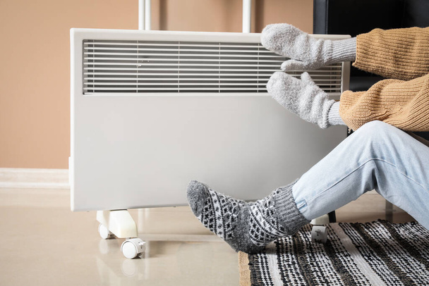 Mujer joven en calcetines calientes y guantes cerca de calentador eléctrico en casa. Concepto de temporada de calefacción - Foto, Imagen