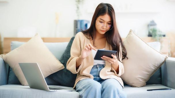 Mujer asiática joven con gafas y el uso de la tableta mientras está sentado en el sofá en casa - Foto, imagen