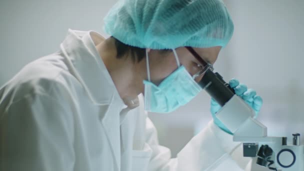 Азіатський вчений носить захисну маску, стерильний капелюх і рукавички з використанням мікроскопа і робить нотатки під час проведення досліджень в лабораторії - Кадри, відео