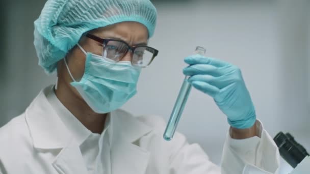 Chest up shot of Asian scientist in protective face mask, medical hat and gloves holding test tube and examining blue chemical in it while working in laboratory - Footage, Video