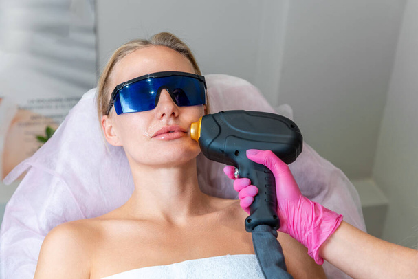 Portrait of young woman at laser hair removal session. Beautician removes hair above client's upper lip. Concept of laser hair removal and aesthetic medicine. - Photo, Image