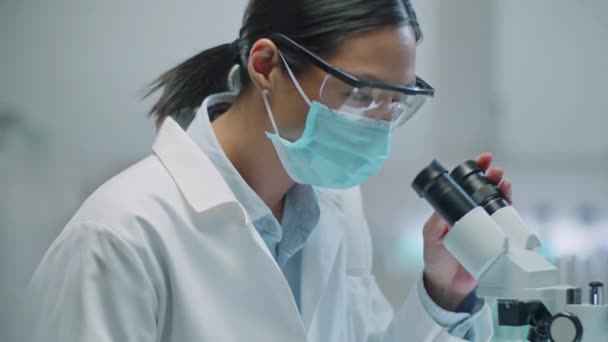 Petto up shot di scienziata asiatica femminile in maschera protettiva e occhiali con microscopio e prendere appunti durante la conduzione di ricerche di laboratorio - Filmati, video
