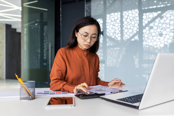 Confident serious concentrated and focused business woman behind paper work inside office, woman with calculator thinking checking accounts financial reports, Asian with laptop preparing documents. - Fotó, kép
