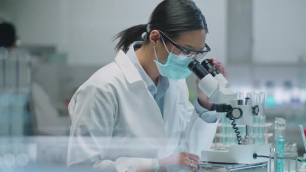 Keskikokoinen laukaus naispuolisesta aasialaisesta tiedemiehestä suojakasvonaamiossa ja turvalaseissa mikroskoopilla ja kirjaamalla ylös tutkimustulokset laboratoriossa - Materiaali, video
