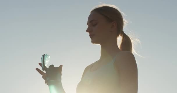 Jeune athlète femme boit de l'eau de bouteille après une séance de jogging en plein air. Femme en vêtements de remise en forme repose après la course et reconstituer la soif - Séquence, vidéo