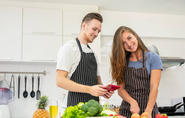 Cheerful young couple lover cooking  together according to a recipe on tablet computer. Smiling woman cuts vegetables, husbund looks at tablet in kitchen interior with vegetables on table and glass of wine. - Photo, Image
