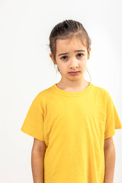 Triest blank klein meisje in een geel T-shirt op een witte achtergrond geïsoleerd, portret van een beledigd kind. - Foto, afbeelding