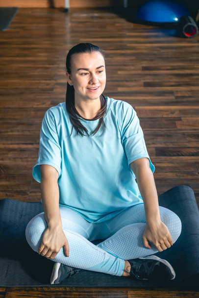Spor kıyafetli genç bir kadın spor salonunda mindere oturmuş yoga yapıyor ve esneme hareketleri yapıyor.. - Fotoğraf, Görsel