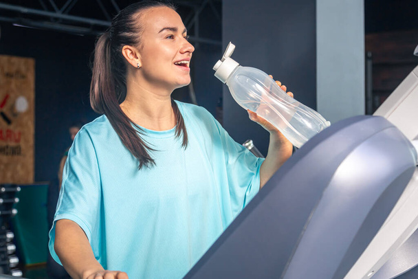 Kobieta pije wodę na siłowni stojąc na bieżni po treningu wytrzymałościowym. - Zdjęcie, obraz