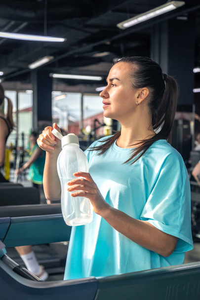 Uma mulher bebe água no ginásio enquanto está em uma esteira após um treino de cardio. - Foto, Imagem