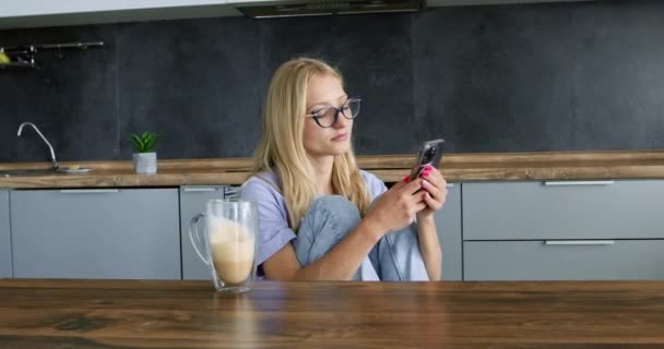 Jonge vrouw met mobiele telefoon drinkt cappuccino in de keuken. Lady leest social media nieuwsfeed rustend met koffie thuis. Vrije tijd en vrije tijd - Video