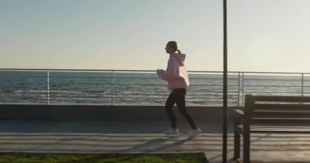 Mujer en chándal corre a lo largo de la ciudad frente al mar en la mañana soleada. Dama delgada disfruta trotando en terraplén vacío. Entrenamiento y ejercicio regular - Imágenes, Vídeo