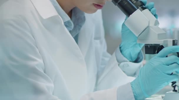 Tilt down shot of female Asian scientist in lab coat and medical gloves using compound microscope and writing down notes while conducting research in lab - Metraje, vídeo