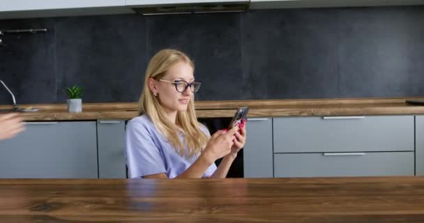 Nuori blondi nainen istuu keittiössä mobiili gadget. Lady käyttää kännykkäverkkoa viihteenä. Hyvä lukea viestejä ja nautintoa ilo - Materiaali, video