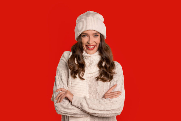 Mladá žena v zimě pletená čepice a svetr pózuje s veselým výrazem, připravená na prázdninové oslavy na červeném pozadí studia, s důvěrou zkřížené ruce a s úsměvem - Fotografie, Obrázek