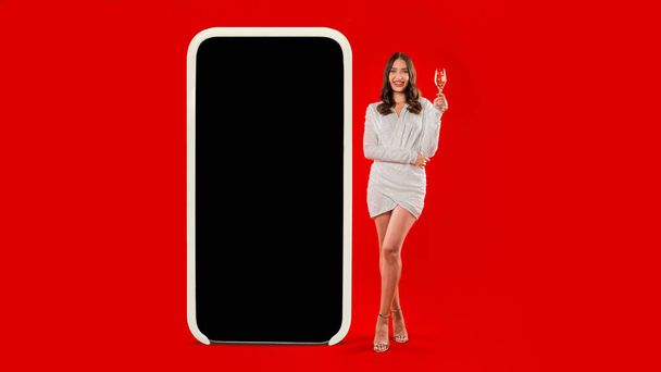 Jonge blanke dame in elegante jurk staat in de buurt van een enorme telefoon met blanco scherm mockup, toast met glas mousserende wijn, tegen rode studio achtergrond. Volledige lengte, panorama - Foto, afbeelding