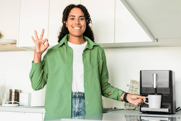 Smiling Black Young Lady Making Coffee Via Machine And Gesturing Okay To Camera In Modern Home Kitchen Interior. Woman Approving Coffee Maker With Ok Sign In The Morning - 写真・画像