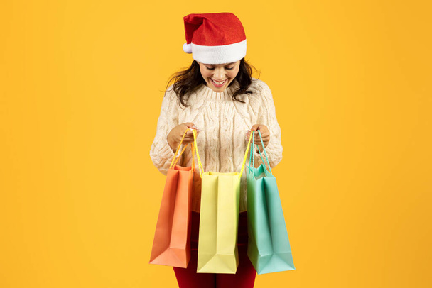 Młoda kobieta ściskając kolorowe papierowe torby na zakupy w kapeluszu Mikołaja, emanuje radością na żółtym tle studio, uosabiając świątecznego ducha handlu Bożego Narodzenia i sezonu sprzedaży - Zdjęcie, obraz
