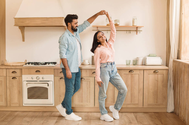 Happy hindu couple enjoys playful dance in their kitchen, filled with natural light and featuring wooden cabinets, embodying joyful domestic life moment - Fotoğraf, Görsel