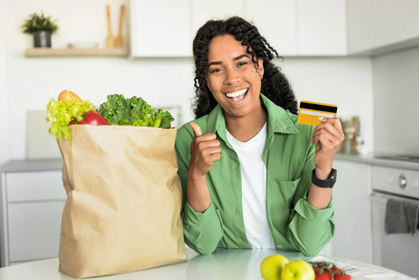 Jonge Afro-Amerikaanse dame met papieren boodschappentas met kredietkaart, duimen opgevend ter goedkeuring van de bezorgdienst, zittend aan tafel in haar thuiskeuken. Financiële kennis - Foto, afbeelding
