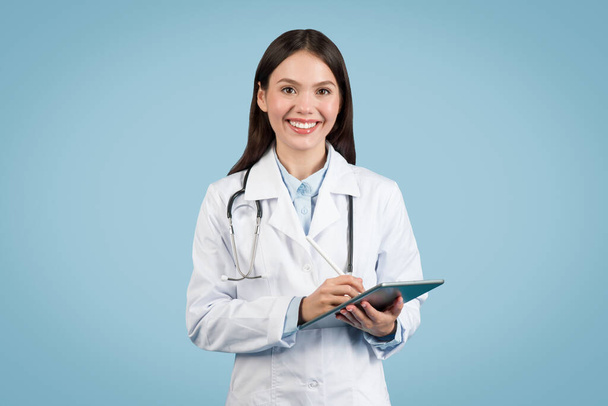 Щаслива, доступна жінка-лікар у білому пальто зі стетоскопом, тримає цифровий планшет, стоїть на синьому фоні і посміхається на камеру - Фото, зображення