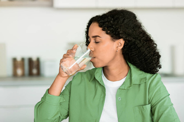 Czarna kobieta cieszy się orzeźwiającą szklanką wody w swojej kuchni w pomieszczeniach, odzwierciedlając zdrowy styl życia, nawilżenie i zrównoważonej diety dla zdrowia odchudzania. Afroamerykanka popija wodę mineralną - Zdjęcie, obraz