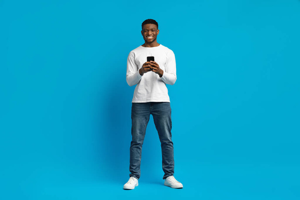 青いスタジオの背景にモダンな携帯電話を使用してカジュアルな服装でポジティブ笑顔の幸せな若いアフリカ系アメリカ人男性,最新のモバイルアプリをチェック,スペースをコピー,全長ショット - 写真・画像