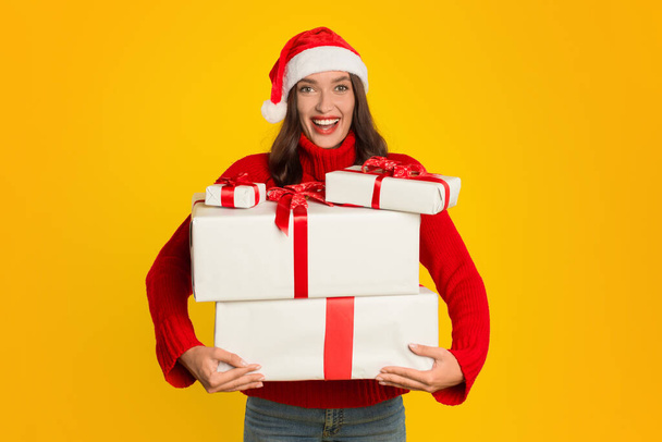 Young lady in Santa hat holds many wrapped Xmas gifts, joyfully congratulating smiling to camera, on yellow studio background. Christmas promotions banner, celebration and gifting concept - Photo, Image