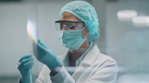 Asian female scientist in protective face mask, disposable hat and gloves adding blue chemical with pipette into test tube and mixing substance during laboratory research - Footage, Video