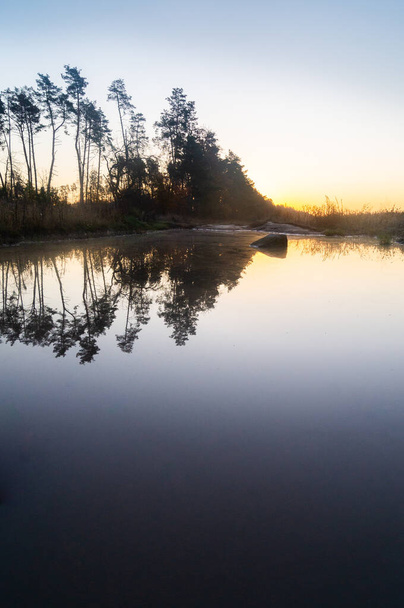 Die goldenen Farbtöne der Morgendämmerung spiegeln sich auf einem stillen Teich wider; die ruhige ukrainische Landschaft spiegelt sich in ruhigen Gewässern wider; der sanfte Sonnenaufgang leuchtet über der ruhigen Wasserstraße.  - Foto, Bild