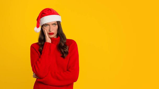 European woman with red Santa hat poses with displeased expression against yellow studio backdrop, embodying expressing disappointment and stress of seasonal holiday shopping. Panorama, empty space - Photo, Image