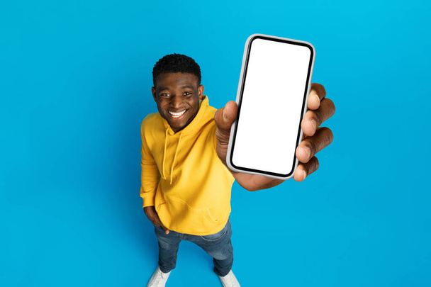 Nagyszerű ajánlat, legújabb mobil alkalmazás, izgalmas online ajánlat. Cool fiatal afro-amerikai srác bemutató telefon fehér üres mockup képernyő, elszigetelt kék stúdió háttér, nagy látószögű nézet - Fotó, kép