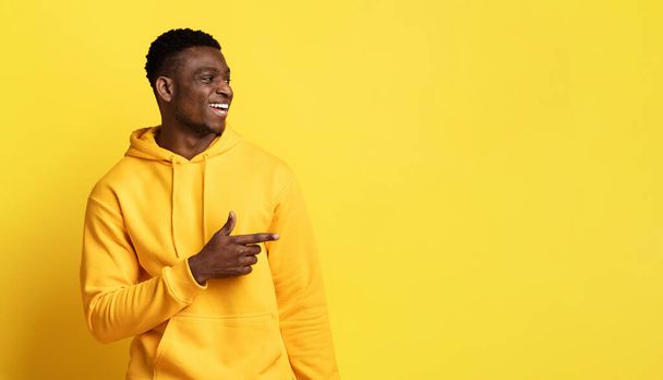 Cheerful handsome young black guy wearing yellow hoodie pointing at copy space and smiling, checking nice offer, advertisement, deal on yellow background, panorama - Foto, Bild