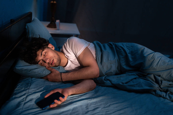 Homme arabe couché sur un oreiller confortable avec un téléphone portable à la main et dormant sous la couverture la nuit. Concept de dépendance et d'insomnie - Photo, image