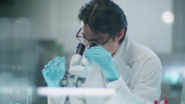 Mies Aasian tiedemies lab takki ja suojakäsineet läpi mikroskoopin ja kirjoittaa muistiinpanoja kokeilun aikana laboratoriossa - Materiaali, video