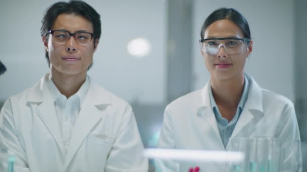 Ritratto di chimici asiatici di sesso maschile e femminile in camici bianchi e occhiali che posano insieme per la macchina fotografica sul posto di lavoro in laboratorio - Filmati, video