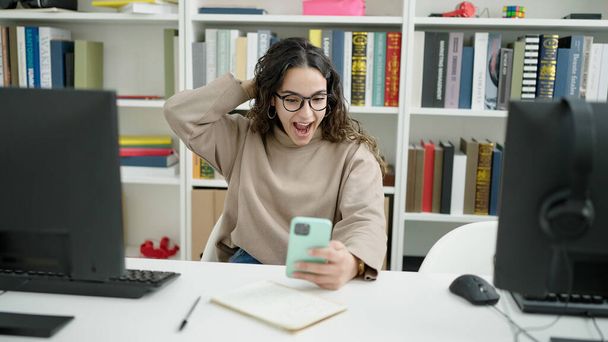 Junge schöne hispanische Studentin benutzt Smartphone mit überraschendem Gesichtsausdruck in der Bibliothek Universität - Foto, Bild