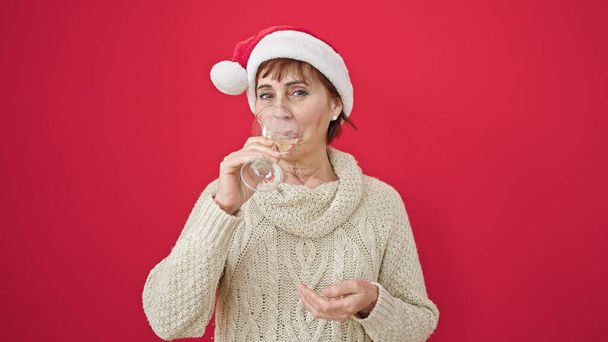 Mulher hispânica madura sorrindo confiante vestindo chapéu de Natal bebendo um copo de vinho branco sobre fundo vermelho isolado - Foto, Imagem