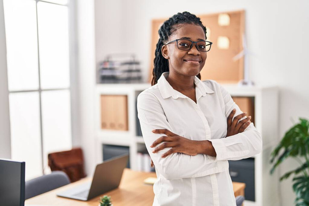 Trabajadora de negocios afroamericana sonriendo confiada de pie con los brazos cruzados gesto en la oficina - Foto, Imagen