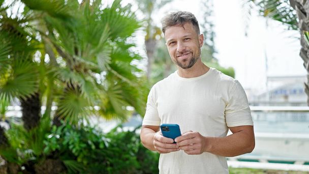 Nuori mies käyttää älypuhelinta hymyillen puistossa - Valokuva, kuva