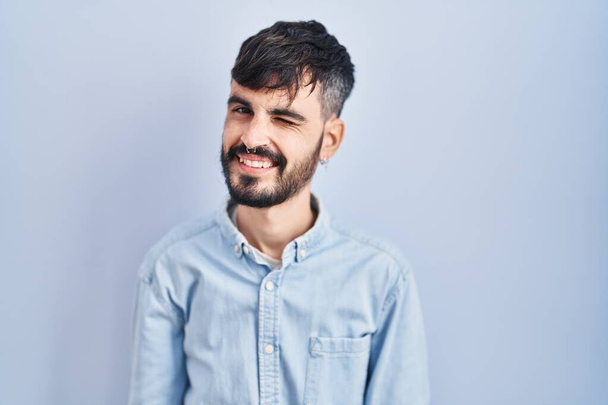 Mladý hispánec s vousy stojící nad modrým pozadím mrkající na kameru se sexy výrazem, veselý a šťastný obličej.  - Fotografie, Obrázek