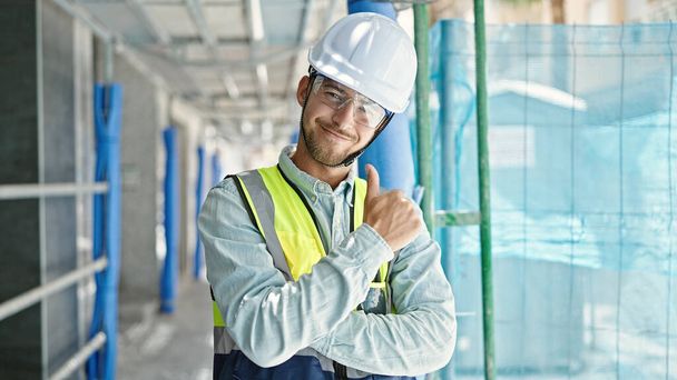 Jeune homme caucasien architecte debout avec les bras croisés geste faire pouce vers le haut geste à la place de la construction - Photo, image