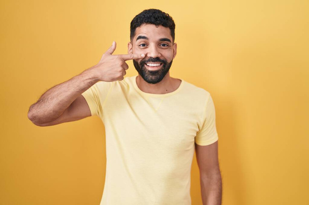 Hombre hispano con barba de pie sobre fondo amarillo señalando con la mano el dedo a la cara y la nariz, sonriendo alegre. concepto de belleza  - Foto, Imagen