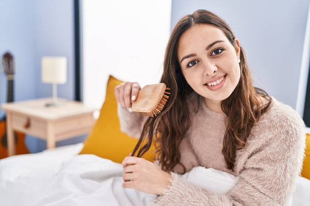 Молода красива іспаномовна жінка розчісує волосся, сидячи на ліжку в спальні
 - Фото, зображення