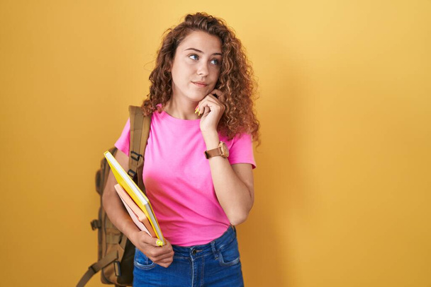 Νεαρή καυκάσια γυναίκα που φοράει φοιτητικό σακίδιο και κρατάει βιβλία με το χέρι στο πηγούνι σκεπτόμενη την ερώτηση, την σκεπτική έκφραση. Χαμογελώντας με ευγενικό πρόσωπο. έννοια της αμφιβολίας.  - Φωτογραφία, εικόνα