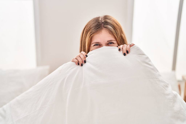 Jeune femme blonde couvrant la bouche avec une couverture assise sur le lit souriant à la chambre - Photo, image