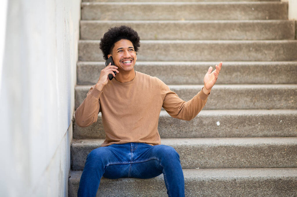Portret gelukkige jonge Spaanse man zit op de trap te praten met telefoon - Foto, afbeelding