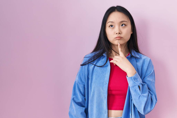 Nuori aasialainen nainen seisoo vaaleanpunainen tausta ajattelu keskittynyt epäilystä sormella leuka ja etsivät ylös ihmettelevät  - Valokuva, kuva